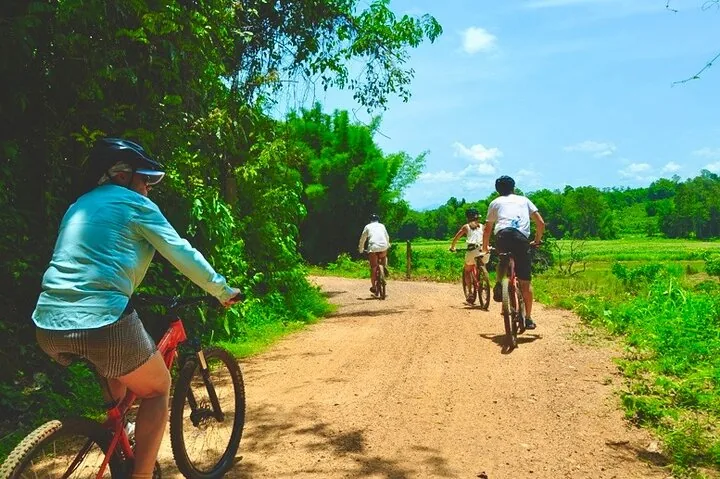 Cyclists riding on a sandy dirt road through a rural area with lush greenery.